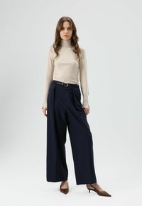 Pull à col roulé beige à manches longues avec des détails de boutons, associé à un pantalon large bleu marine et des escarpins marron à bouts pointus.