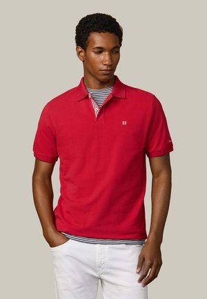 Joven de pie vistiendo una camiseta polo roja sobre una camiseta de rayas, pantalones blancos y zapatos negros sin cordones, frente a un fondo beige liso.