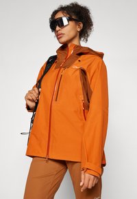 Orange waterproof jacket with a brown hood and zippers, featuring a chest pocket and adjustable cuffs. Model wears black sunglasses.