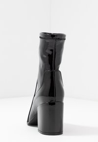 Botín negro de charol con un tacón cuadrado, textura suave y detalle de costura contrastante en la parte superior. Diseño y forma minimalista.