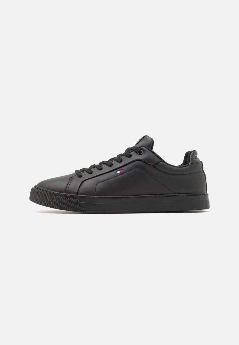 Baskets basses noires avec lacets, semelle texturée et petit logo rouge, blanc et bleu sur le côté, sur fond blanc uni.