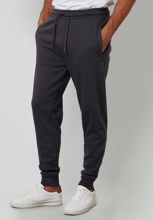 Pantalones de chándal gris oscuro con cintura elástica y cordón, que cuentan con bolsillos laterales y puños ajustados, combinados con zapatillas blancas.