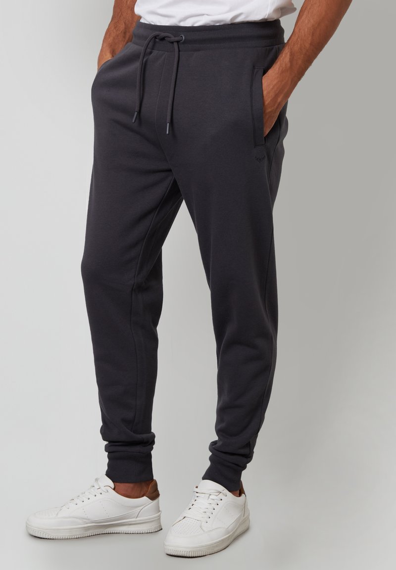 Mørkegrå sweatpants med elastisk talje og snøre, med sidelommer og tapered manchetter, parret med hvide sneakers.