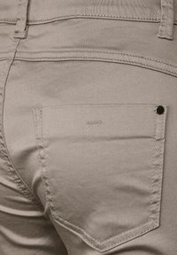 Ljusbeiga byxor med en slät yta och synliga sömmar. Har en bakficka med en svart metallnitar och en subtil logotyp.