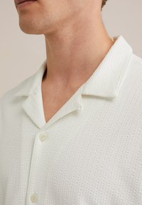 Camicia bianca leggera con un pattern testurizzato, colletto a punta e chiusura con bottoni. Il tessuto sembra traspirante e leggermente strutturato.