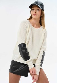 Cream ribbed knit sweater with black leather sleeves. Paired with a black leather mini skirt. Model wears a black cap, showcasing a casual look.