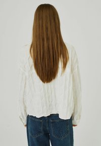 Vit, strukturerad blus med en vågig design, långa ärmar och en kortad nederkant, kombinerad med högmidjade blå denimjeans.