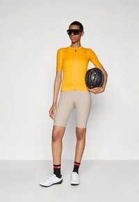 Ποδηλάτης με πορτοκαλί φανέλα, μπεζ σορτς και μαύρα γυαλιά ηλίου κρατάει ένα μαύρο κράνος, φορώντας λευκά παπούτσια ποδηλάτου και μαύρες κάλτσες.