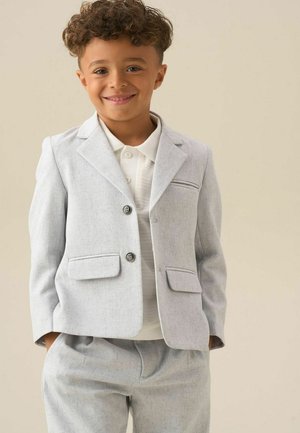 Lachende jonge jongen met krullend haar draagt een lichtgrijs colbert en bijpassende broek over een wit poloshirt, handen in de zakken.