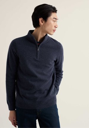 Tmavomodrý pulóver z mäkkej látky s stojatým golierom a štvrťzipsom, nosený cez pásikavú košeľu, doplnený tmavými džínsami.