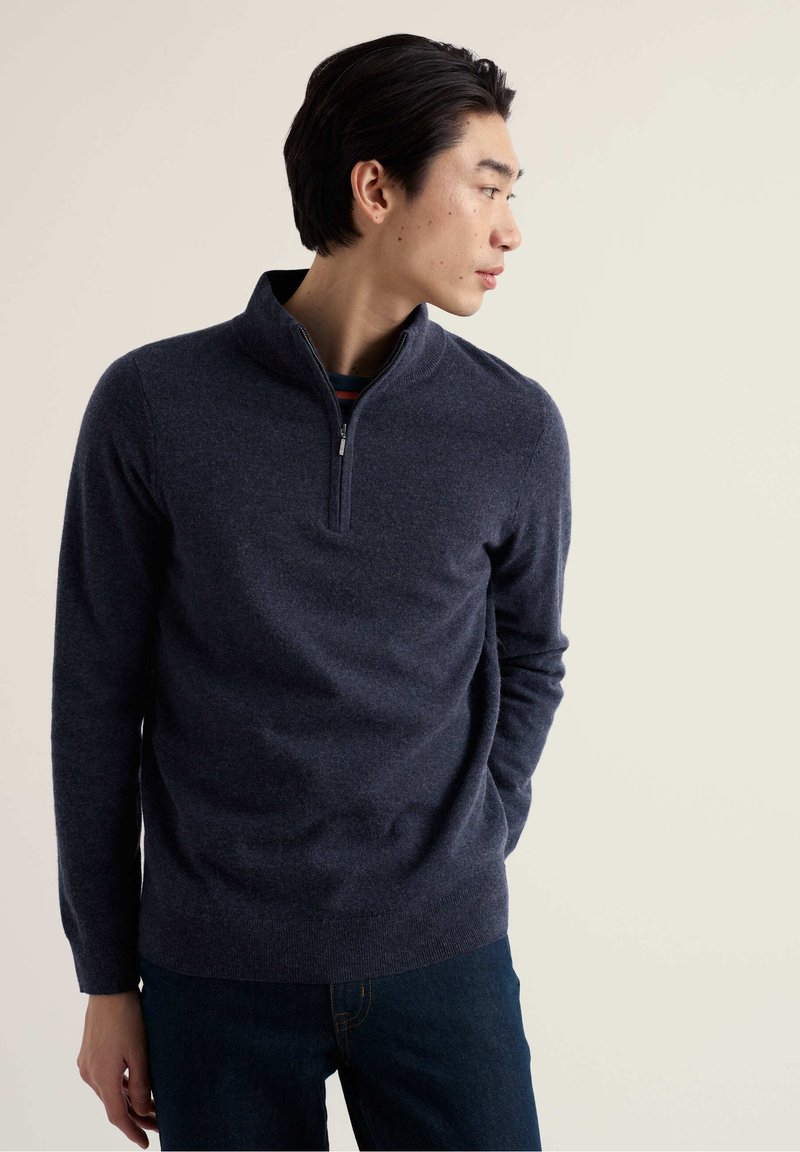 Tumesinist värvi pehme kangaga pullover, millel on seisev krae ja veerandlukustus, kantud triibulise särke peal, kombineeritud tumedate teksadega.