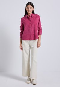 Camicia button-up in corduroy rosa con maniche lunghe, abbinata a pantaloni larghi bianco sporco e sneaker bianche. Design minimalista e tessuto texturizzato.