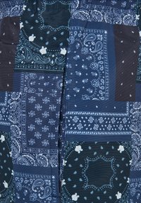 Pamut bandana mintás szövet, amely különböző kék árnyalatokkal és fehér paisley mintákkal, valamint kontrasztos sötét foltokkal rendelkezik. Rugalmas derékrész részlet.