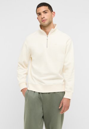 Man wearing a cream quarter-zip pullover sweater and olive green pants, standing with one hand in pocket against a plain background.