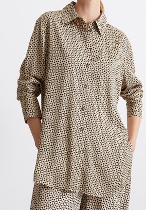 Beige and black geometric patterned long-sleeve button-up shirt with collar, worn with matching pants, hands in pockets.