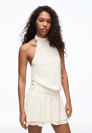 Kobieta o długich, kręconych włosach, ubrana w bezrękawnikową, białą sukienkę o wysokim dekolcie z fakturowanego materiału, z rozkloszowanym dołem na białym tle.