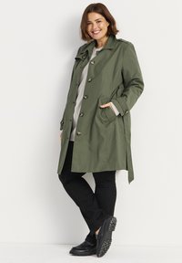 Olijfgroene trenchcoat met een ceintuur in de taille, grote knopen en zijzakken, gedragen over een lichtgrijs topje en zwarte broek. Zwarte schoenen met ribbelzolen.