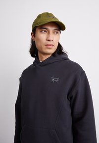 Felpa nera con tasca frontale, con un piccolo logo Reebok grigio. Un cappellino verde con un logo è posizionato sulla testa di un modello.