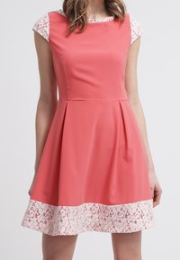 Bezrękawna sukienka w kolorze koralowym z białą koronką na dole i białymi koronkowymi krótkimi rękawami pod spodem.
