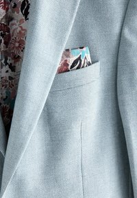 Lyseblå blazer med en tekstureret stof; har en blomsterlignende lommetørklæde med farver som pink, blå og sort, der stikker frem fra lommen.