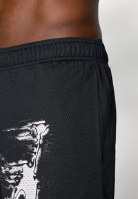 Sidebillede af en person iført sorte mesh shorts med et hvidt grafisk design på venstre lår og en elastisk talje.