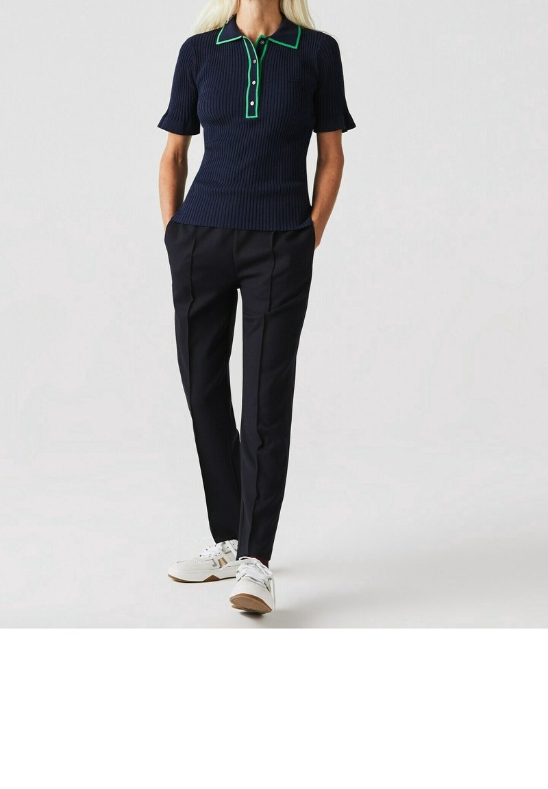 Femme portant un polo côtelé bleu marine à manches courtes avec bordure verte, un pantalon noir sur mesure et des baskets blanches, debout les mains dans les poches.