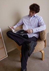 Wit en blauw gestreept overhemde, zwarte gestreepte broek, zwarte schoenen, zittend op een gestructureerde beige stoel, een tijdschrift lezend.