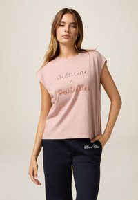 Rosa Baumwoll-T-Shirt mit kurzen Ärmeln, das mit einem strukturierten, metallischen Dekordruck versehen ist. Kombiniert mit navyblauen Hosen und einem Detail in der Seite.