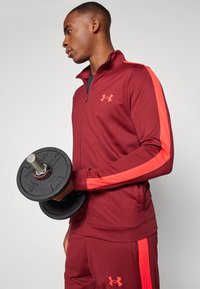 Veste de sport zippée bordeaux avec des rayures orange sur les côtés, associée à un pantalon assorti. La personne tient une plaque de poids noire.