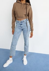 Brun kort hoodie med framficka, ljusblå jeans med hög midja och vita sneakers. Modell står på en blå matta.