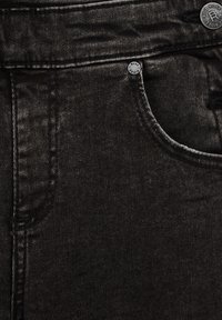 Fekete denim farmernadrág sima textúrával, elülső zsebbel és fém gombokkal. Az anyag színárnyalatain finom eltérések vannak.