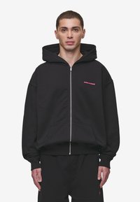 Black zip-up hoodie with a large hood, front pockets, and ribbed cuffs. Features a small pink "PEGADOR" logo on the left chest.