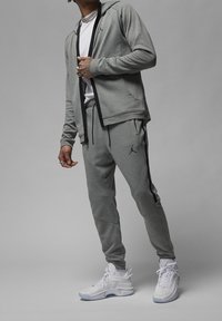 Man in een grijze Jordan trainingspak met zwarte strepen en witte sportschoenen, staande tegen een grijze achtergrond.