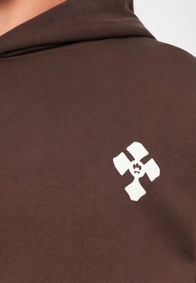 Primo piano di un tessuto marrone con un logo astratto di un fiore a quattro petali bianco stampato vicino all'area del petto.