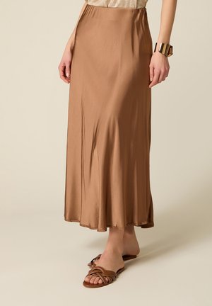 Donna che indossa una lunga gonna marrone di seta con sandali bassi marroni intrecciati e un bracciale rigido d'oro, in piedi su uno sfondo chiaro.