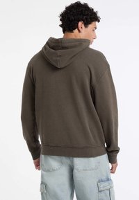Uomo con capelli ricci che indossa un felpa cappuccio marrone scuro e jeans cargo azzurri chiari, di spalle contro uno sfondo bianco semplice.