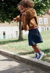 Brun fleecejakke, rødstribet skjorte, denimnederdel, blå og hvide sneakers med solmotiv, hopper på fortovet nær græs og et træ.