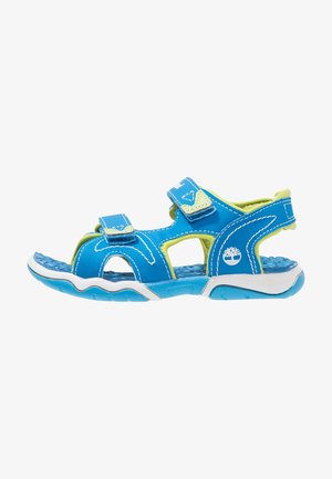 Sandalia azul y verde con correas de Velcro ajustables, diseño abierto, suela texturizada y una tapa de caucho blanca en la puntera y el talón.