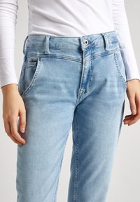 Jeans de denim azul claro con un ligero desvanecido, diseño de cinco bolsillos, pasadores para cinturón y cierre de botón, combinados con una camiseta ajustada de manga larga blanca.