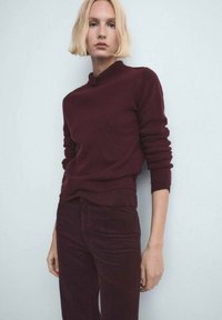 Persoană blondă purtând un pulover burgund închis și pantaloni asortați, stând în fața unui perete de un albastru pal, uitându-se drept înainte.