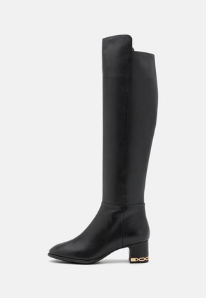 Botte en cuir noir montant jusqu'au genou avec un design épuré, bout pointu, talon carré moyen et accent en chaîne dorée sur le talon. Texture lisse.
