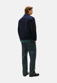 Navy corduroy jas met een kraag, gecombineerd met een groene geruite shirt eronder en losse grijze broek, gedragen met zwarte schoenen.