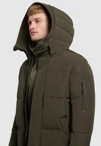 Gewatteerde jas in donkerolijfgroen, met een grote capuchon, een met rits afsluitbare zijzak en horizontale stikseldetails op het lijf.