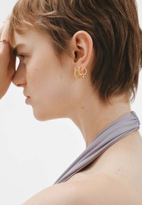 Pendientes de aro dorados con acentos texturizados, llevados por una persona con el cabello corto, que presenta una tela lila drapeada alrededor del cuello.