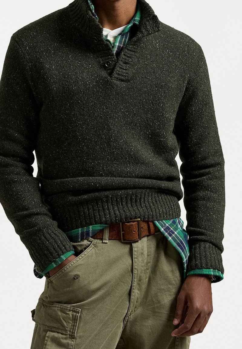 Personne portant un pull en tricot vert foncé par-dessus une chemise à carreaux verte rentrée dans un pantalon cargo kaki avec une ceinture marron.