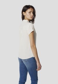 Blusa blanca de manga corta con patrón texturizado y cuello, metida dentro de unos vaqueros azules. La parte trasera presenta un pliegue para mayor detalle.