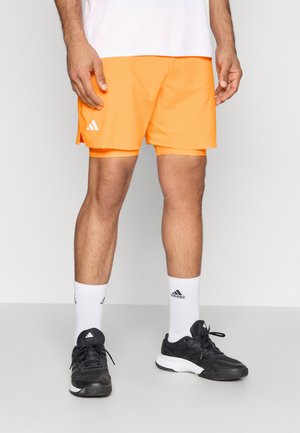 Shorts de sport orange avec des fentes sur les côtés, avec un logo Adidas blanc. Portés avec des chaussures noires et des chaussettes blanches. Texture lisse.