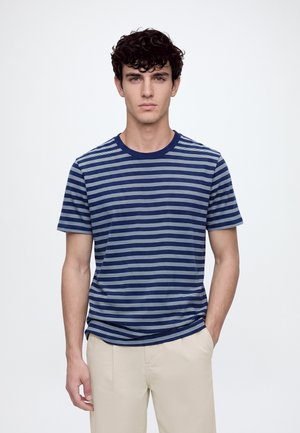 Jeune homme aux cheveux bouclés foncés portant un t-shirt rayé bleu marine et blanc et un pantalon beige clair, debout devant un fond clair uni.