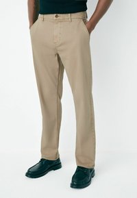 Man wearing beige straight-leg trousers with hands in pockets and black lace-up shoes, standing against a white background.