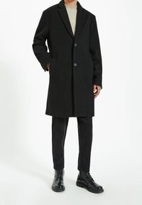 Cappotto di lana nera con rever a punta, due bottoni, tasche laterali, indossato su un maglione di cotone leggero, abbinato a pantaloni neri e stivaletti con lacci.